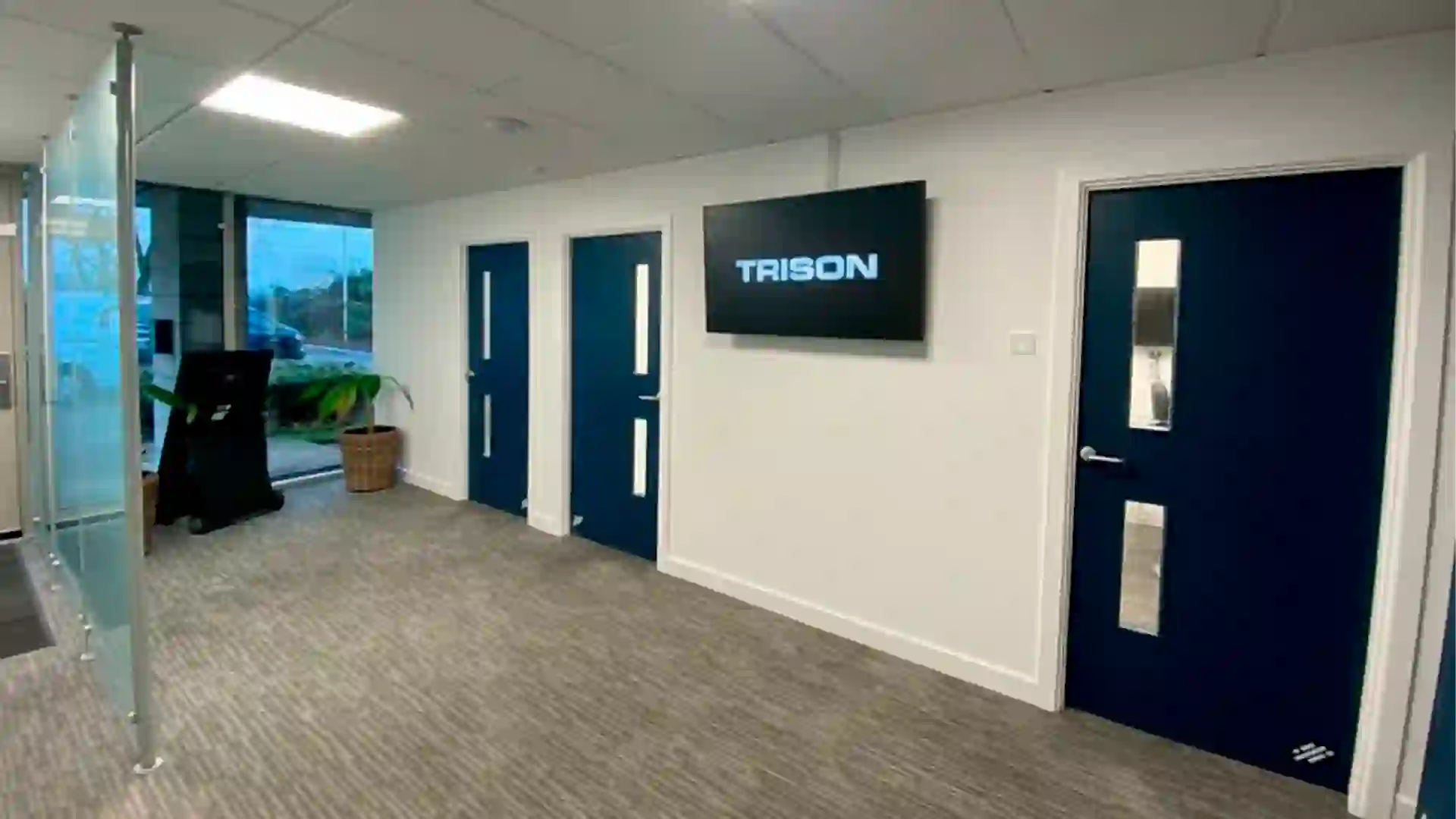 Interior view of the TRISON UK office in Reading, featuring a modern lobby with a digital signage screen displaying the TRISON logo on a white wall, sleek navy blue doors, and minimalist grey flooring.