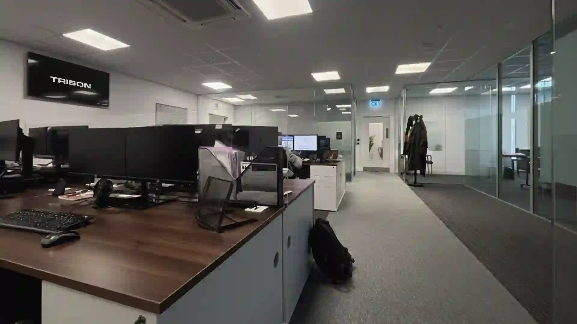 Interior view of the TRISON UK office in Stockport, showcasing a modern open-plan workspace with multiple computer workstations, wooden desks, glass partitions, and a digital screen displaying the TRISON logo.